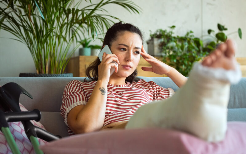 Worried young woman with broken leg in plaster cast lying down on sofa at home and talking on smart phone.