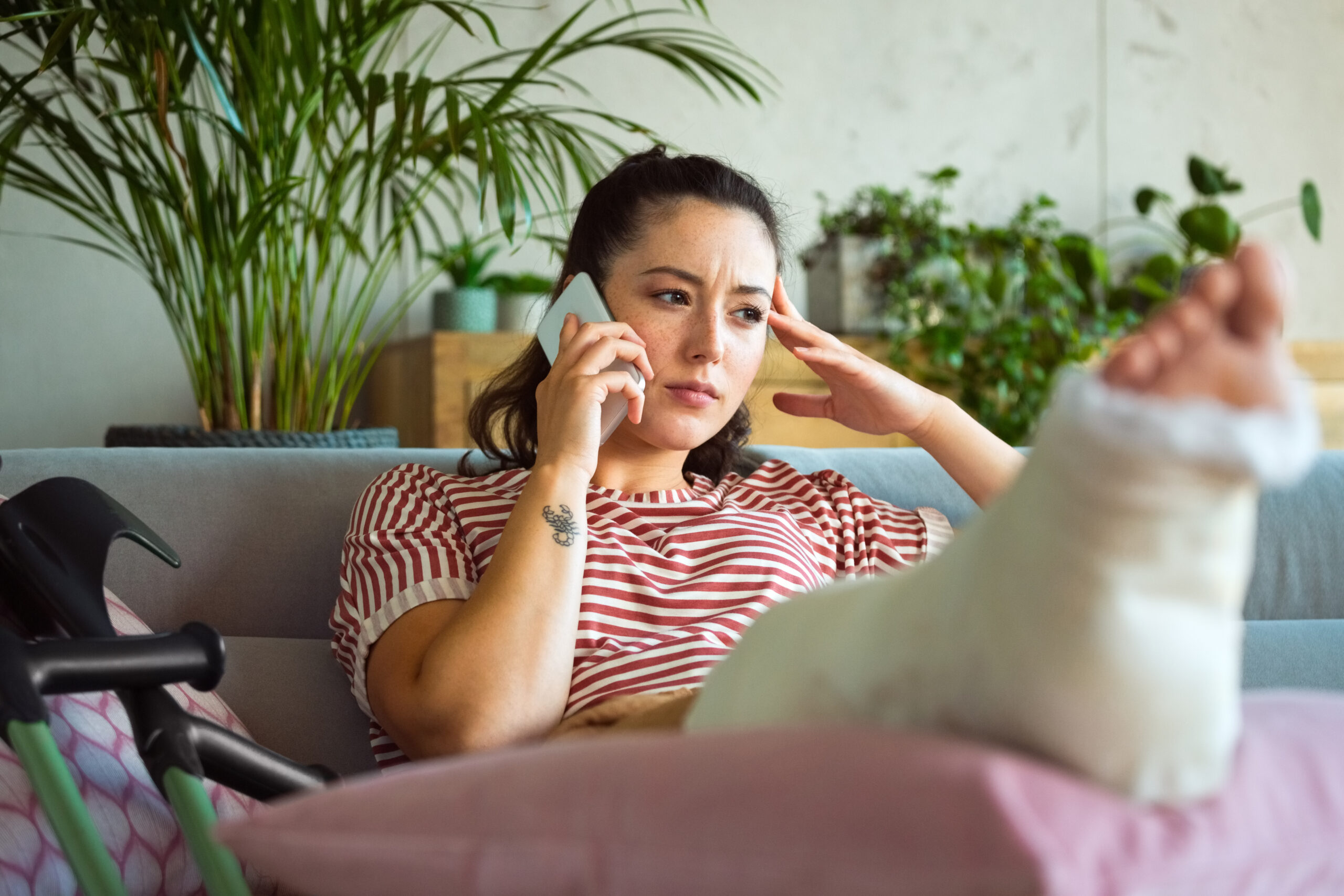 Worried young woman with broken leg in plaster cast lying down on sofa at home and talking on smart phone.