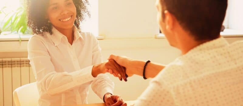 Smiling millennial african woman handshaking partner at office meeting, happy black female manager and satisfied male client or colleague shaking hands making deal or thanking for help in teamwork
