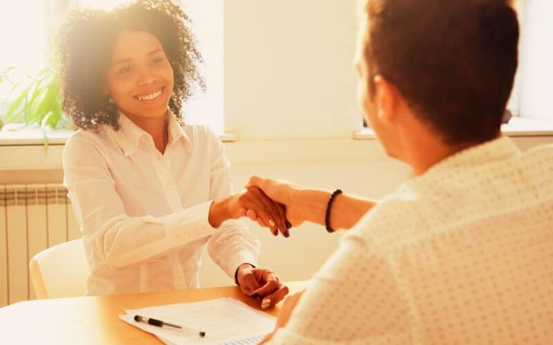 Smiling millennial african woman handshaking partner at office meeting, happy black female manager and satisfied male client or colleague shaking hands making deal or thanking for help in teamwork