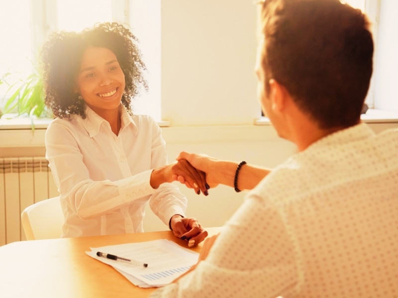 Smiling millennial african woman handshaking partner at office meeting, happy black female manager and satisfied male client or colleague shaking hands making deal or thanking for help in teamwork