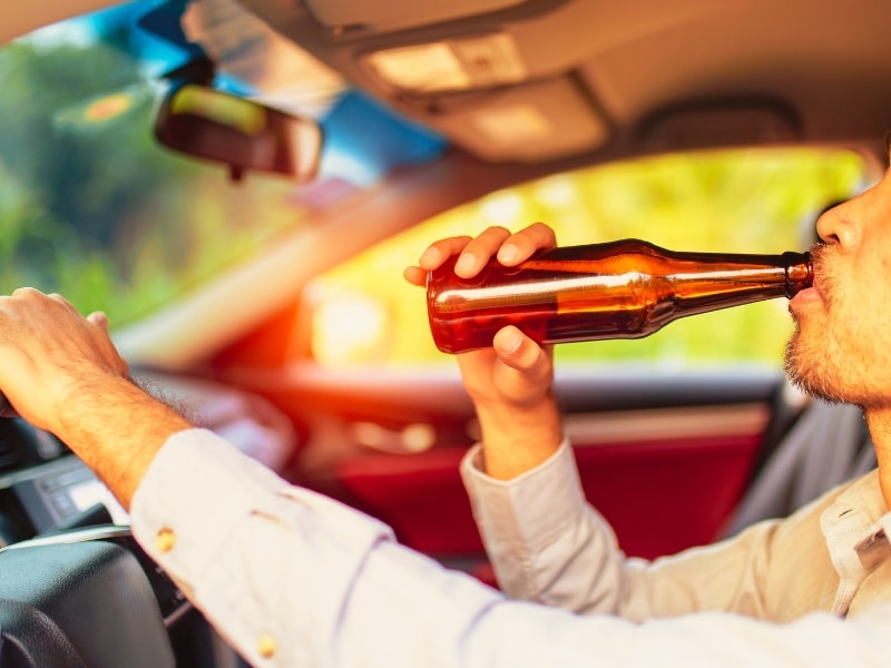 Drunk asian young man drives a car with a bottle of beer with sunset background, Dangerous driving concept