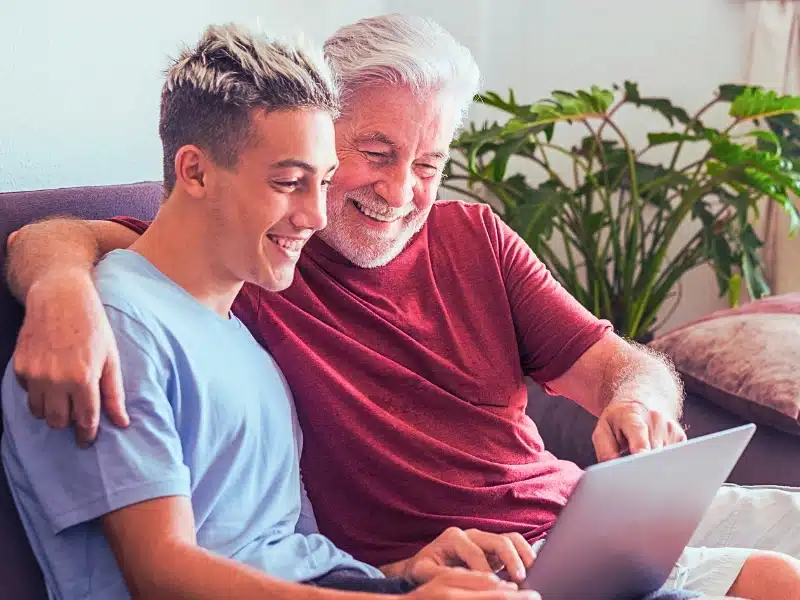 Father and son sitting on the couch looking at the laptop and both smiling.