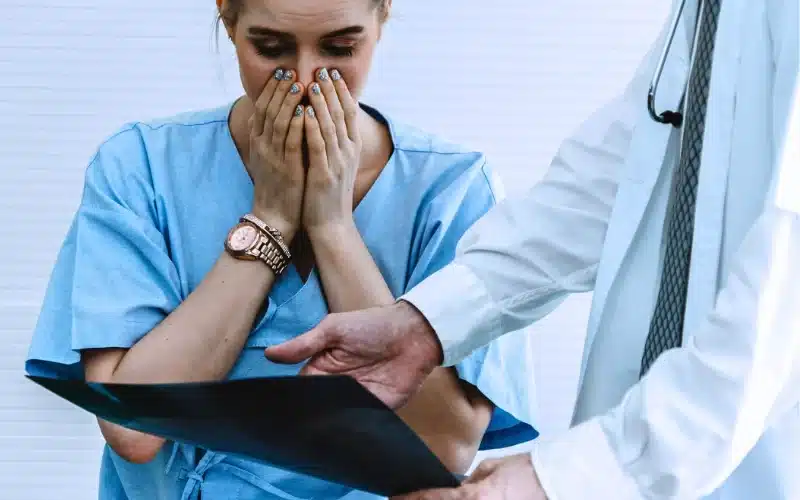 Concerned woman in a medical uniform, covering her mouth while a person in a lab coat points to a document, suggesting a serious or distressing situation.