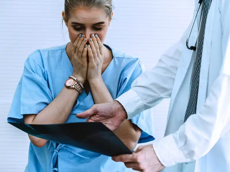 Concerned woman in a medical uniform, covering her mouth while a person in a lab coat points to a document, suggesting a serious or distressing situation.